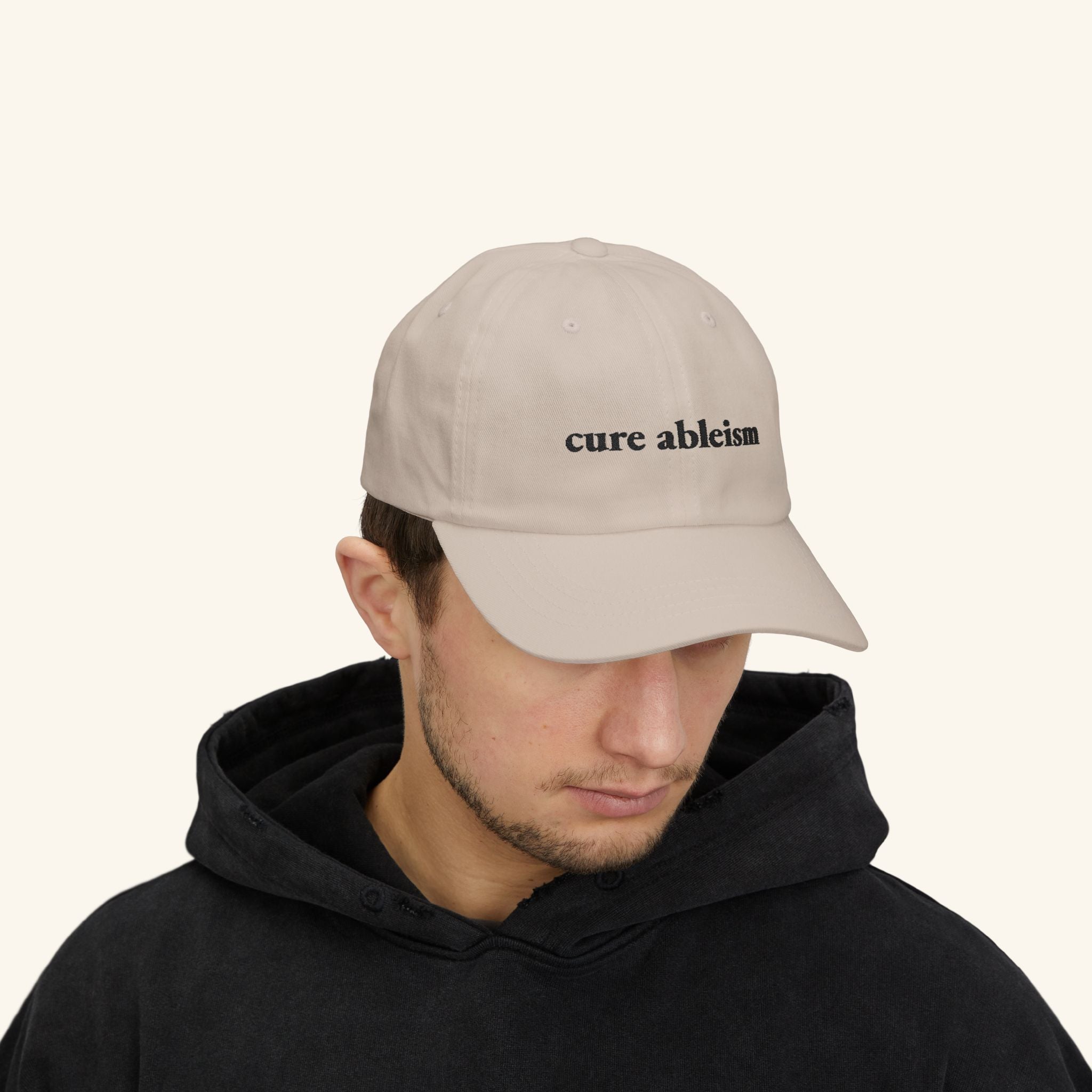 Person wearing a beige cap with 'cure ableism' text on a plain background